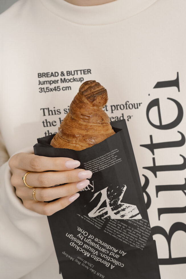 Close-up of a mockup sweatshirt with typography design featuring the words Bread & Butter. A hand holds a croissant in black paper packaging. Fashion design.