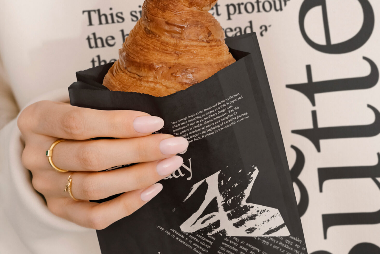 Hand holding croissant in black paper bag with modern typography design. Ideal for graphic designers seeking creative mockup inspiration and unique packaging design.