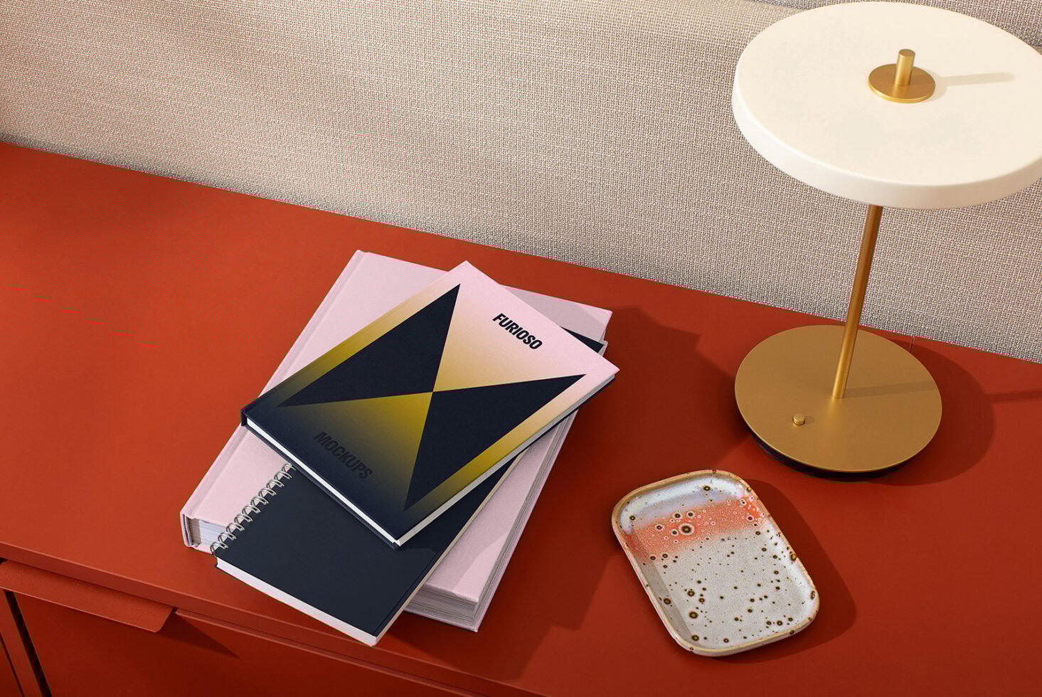 Modern desk setup with stacked notebooks featuring mockup designs, next to a minimalist lamp and ceramic tray on a rust-colored surface.