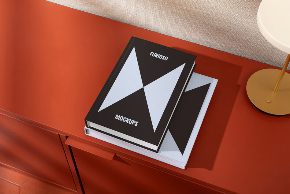Top view of two hardcover book mockups on a red table next to a lamp for designers. Ideal for showcasing graphics templates for creative projects.