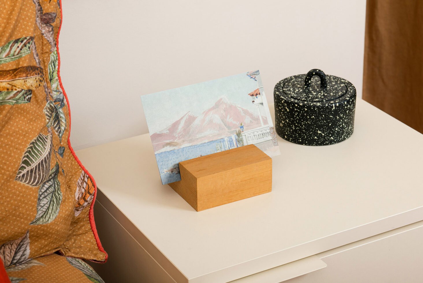 Tabletop mockup featuring a postcard with mountain art in a wooden holder decorative pillow with leaf design and speckled jar perfect for designers.