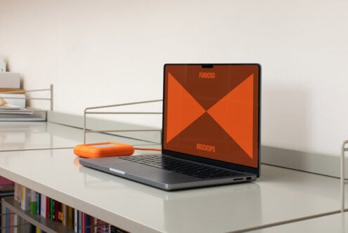 Laptop on desk displaying orange mockup template, adjacent to an orange external drive, ideal for designers seeking modern workspace graphics.