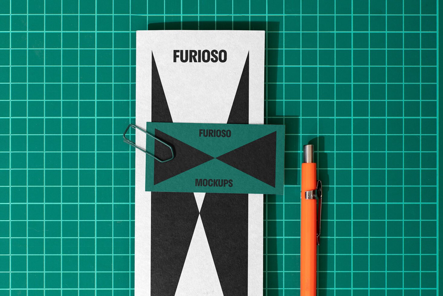 Stationery mockup with business card and paper on a green grid cutting mat. Includes a paper clip and orange pen. Ideal for designers seeking graphics.