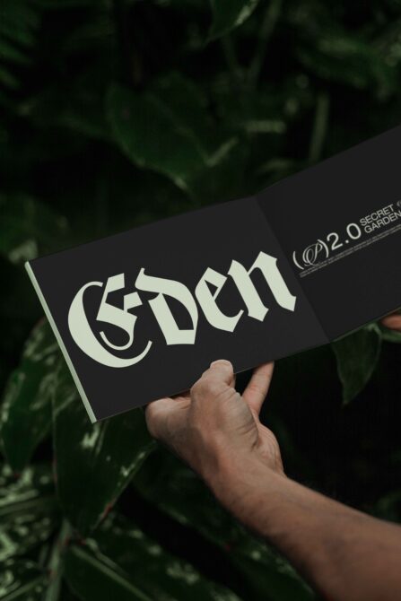 Open book with gothic typography on a black background held by hands against green foliage font design typography mockup for designers print template