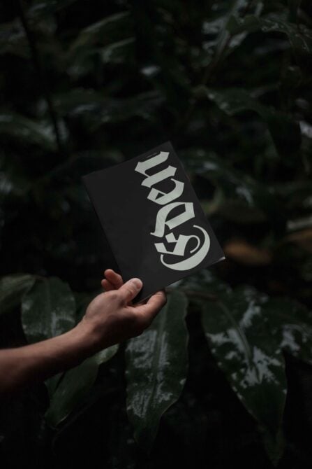 Hand holding a stylish dark booklet with bold font design, surrounded by lush green foliage; perfect for typography and graphic design enthusiasts.