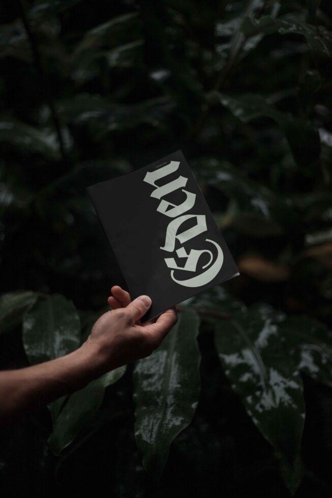 Hand holding a stylish dark booklet with bold font design, surrounded by lush green foliage; perfect for typography and graphic design enthusiasts.