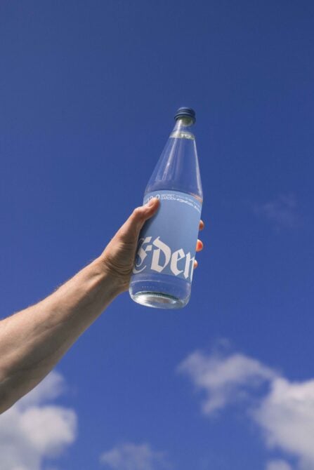 Hand holding glass bottle with blue label against clear sky