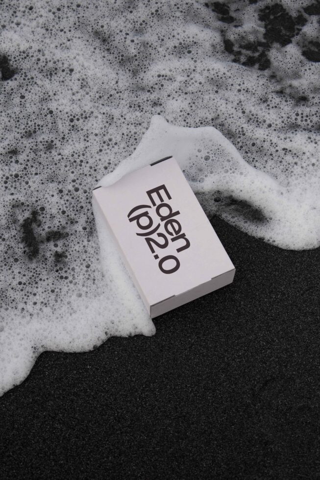 Minimal packaging mockup of a box labeled Eden p2.0 on black sand with ocean foam. Ideal for designers seeking modern branding templates and graphics.