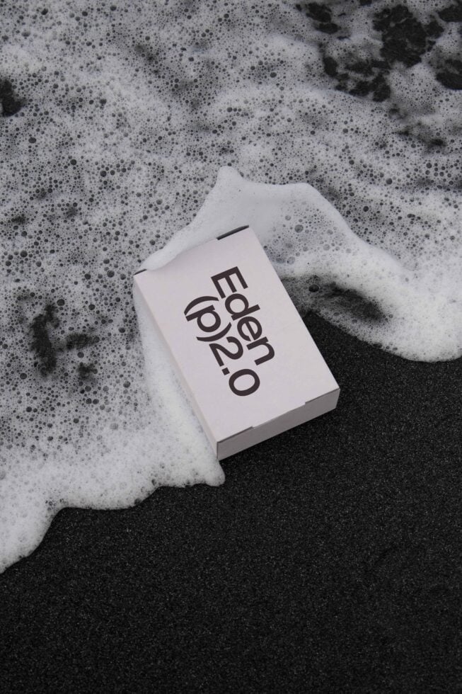 Minimalist mockup of a white box labeled Eden p2.0 on wet sand, surrounded by ocean foam. Ideal for product packaging, branding visuals, and design projects.