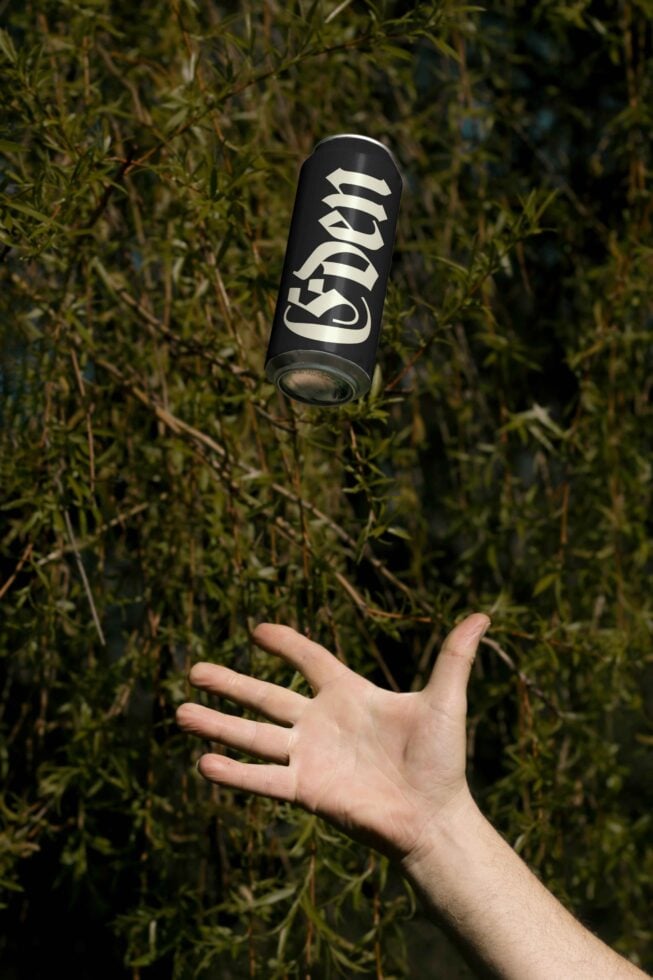 Mockup of a hand tossing a black can with decorative font against a natural leafy backdrop; ideal for product branding and graphic design projects.