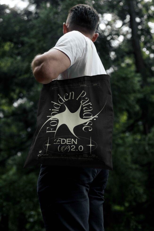 Man holds a tote bag with abstract graphic design Forbidden Paradise text in a forest setting. Ideal for mockup design showcasing unique graphics.
