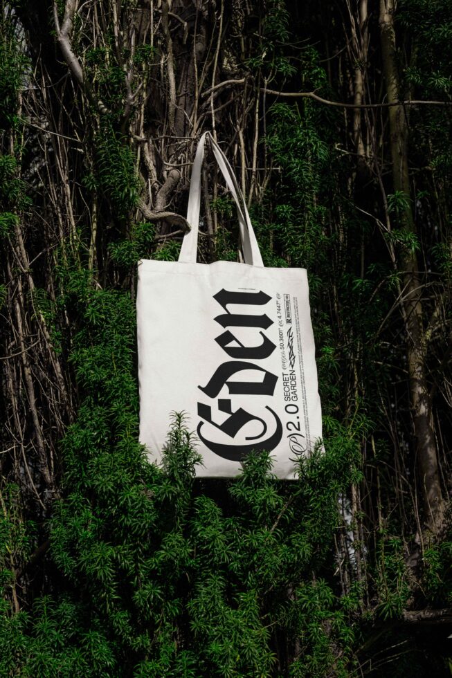 Tote bag with bold typography reading Eden hanging on lush green foliage backdrop natural eco-friendly mockup graphic design asset for creatives.