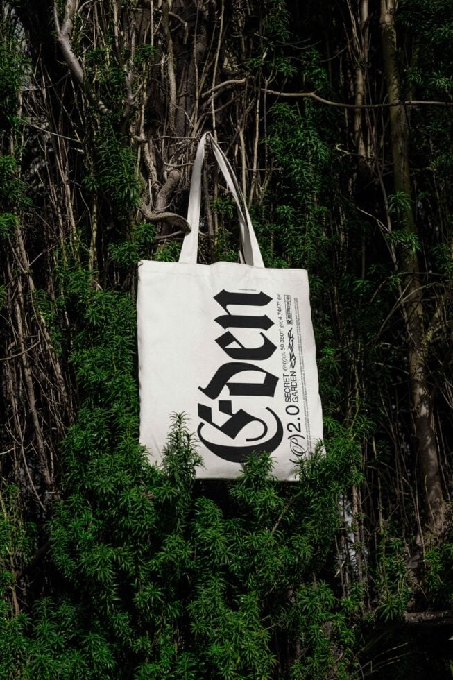 Tote bag mockup with bold typography hanging among lush greenery. Perfect for showcasing branding designs and eco-friendly style. Realistic presentation.