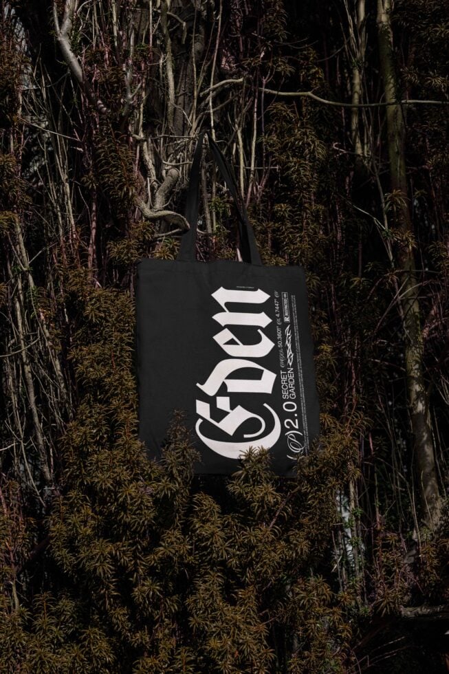 Black tote bag mockup with bold typography design hanging on tree branches in a natural setting perfect for showcasing graphic design assets and templates.