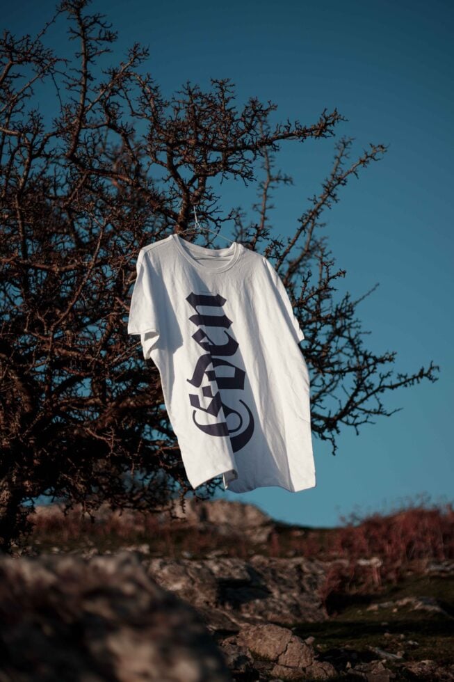White T-shirt with black gothic font hanging from tree branch in natural setting design mockup outdoor photo for designers apparel template SEO keywords