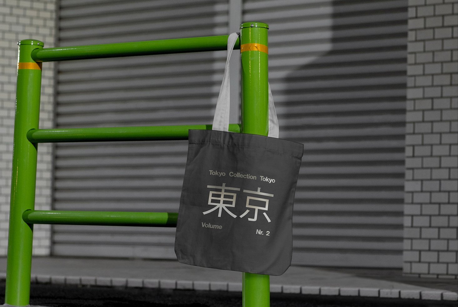 Black tote bag mockup with Tokyo Collection design hanging on a green metal rail. Urban setting with brick wall. Ideal for showcasing graphics.