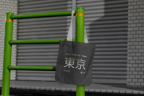 Black tote bag mockup with Tokyo Collection design hanging on a green metal rail. Urban setting with brick wall. Ideal for showcasing graphics.