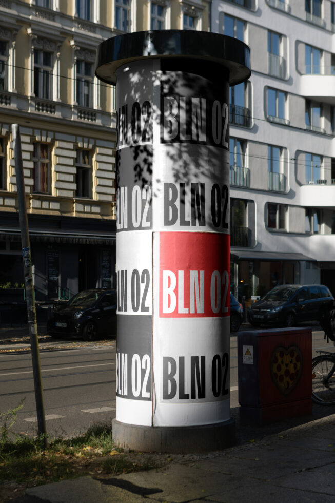 Cylindrical street column wrapped with repeating BLIN02 text in black, white, red bands