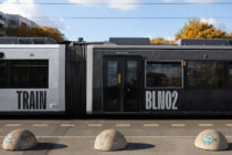 Black train car labeled TRAIN, gray tram labeled BLIN02 with decorated bollards in front