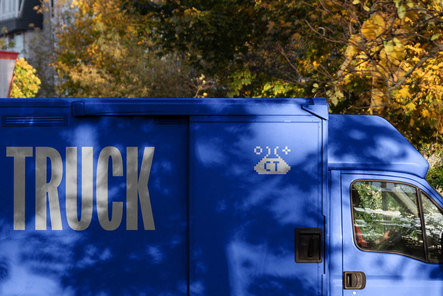 Blue truck with bold TRUCK text and pixel art design on side, set against a backdrop of autumn trees. Ideal for graphic mockup templates.
