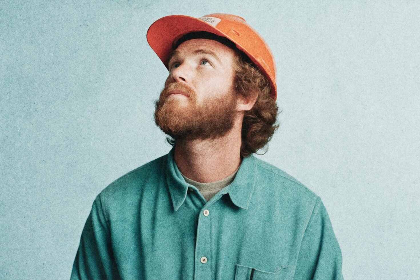 Man in orange hard hat and teal shirt looking upwards. Construction-themed portrait for design assets. Use in graphics or mockups. Rugged style.