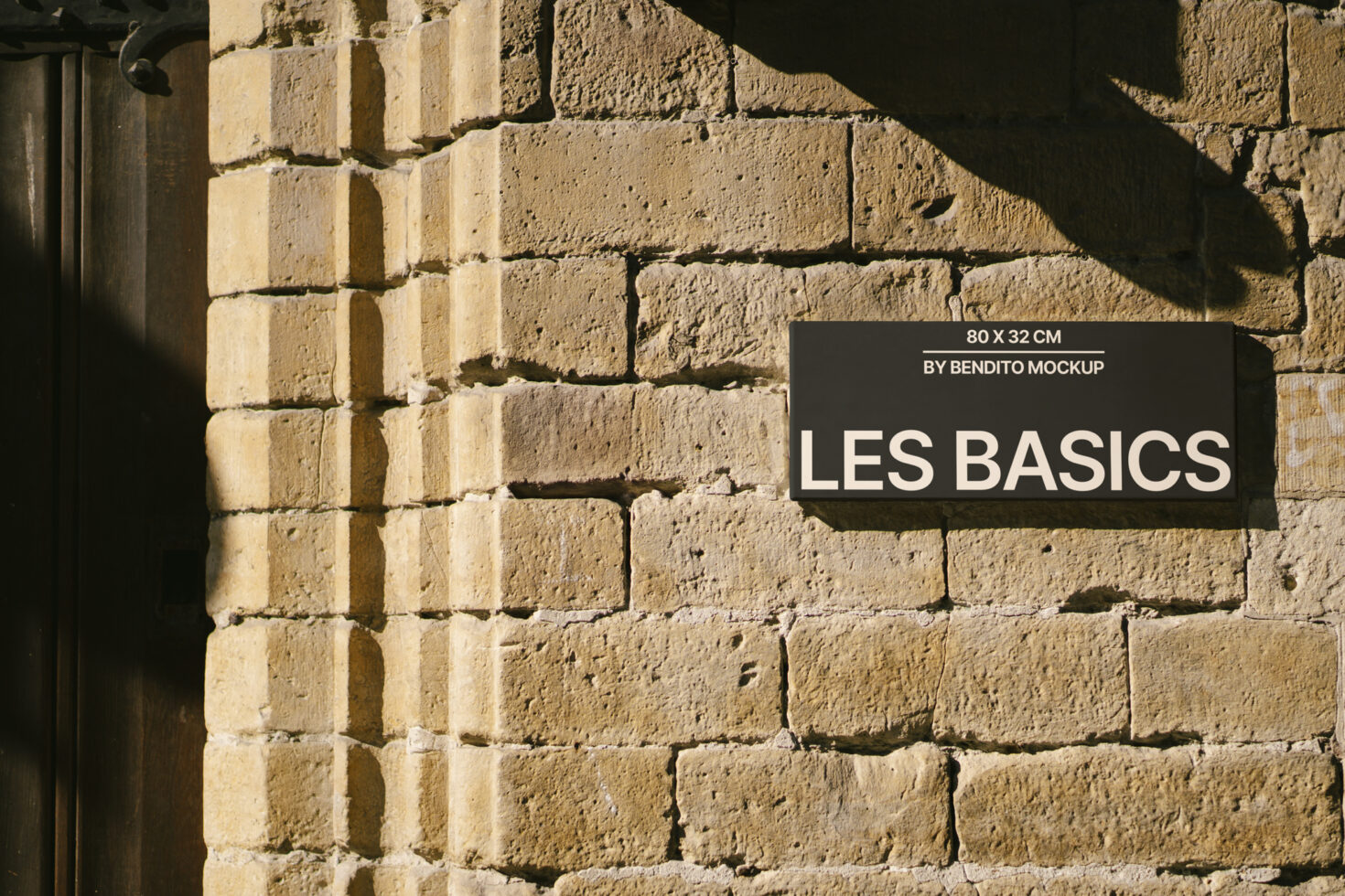 Black rectangular sign with white text on rough sandstone brick wall with dramatic sunlight shadows
