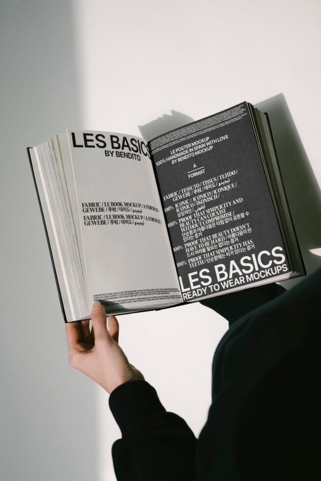 Open book mockup held by a person displaying black and white pages. Design includes text in various languages. Ideal for designers seeking high-quality mockups.