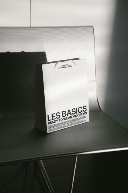 Paper shopping bag mockup with Les Basics typography placed on a modern chair in minimal light. Perfect for designers seeking packaging mockup assets.
