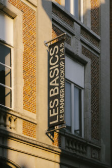 Vertical hanging sign mockup on brick building reads Les Basics Le Banner
