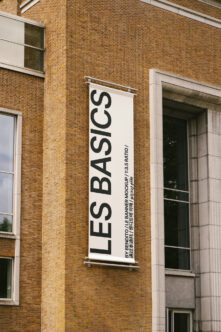 White vertical banner mockup on red brick wall with black text reading LES BASICS