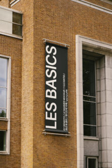 Black vertical banner mockup on red brick facade with white text reading LES BASICS