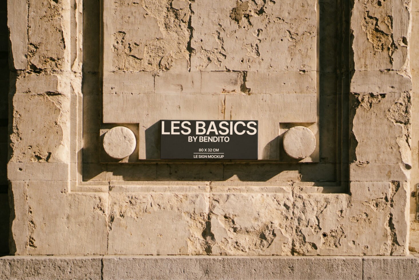 Stone textured wall showcasing a rectangular black sign with bold white text reading Les Basics by Bendito ideal for mockup presentations and design use