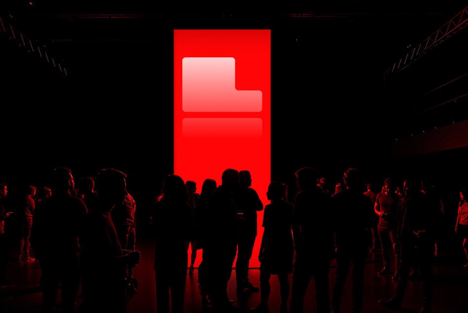 Silhouettes of people stand in front of a large red illuminated screen in a dark room, perfect for technology event graphics, digital art backgrounds.
