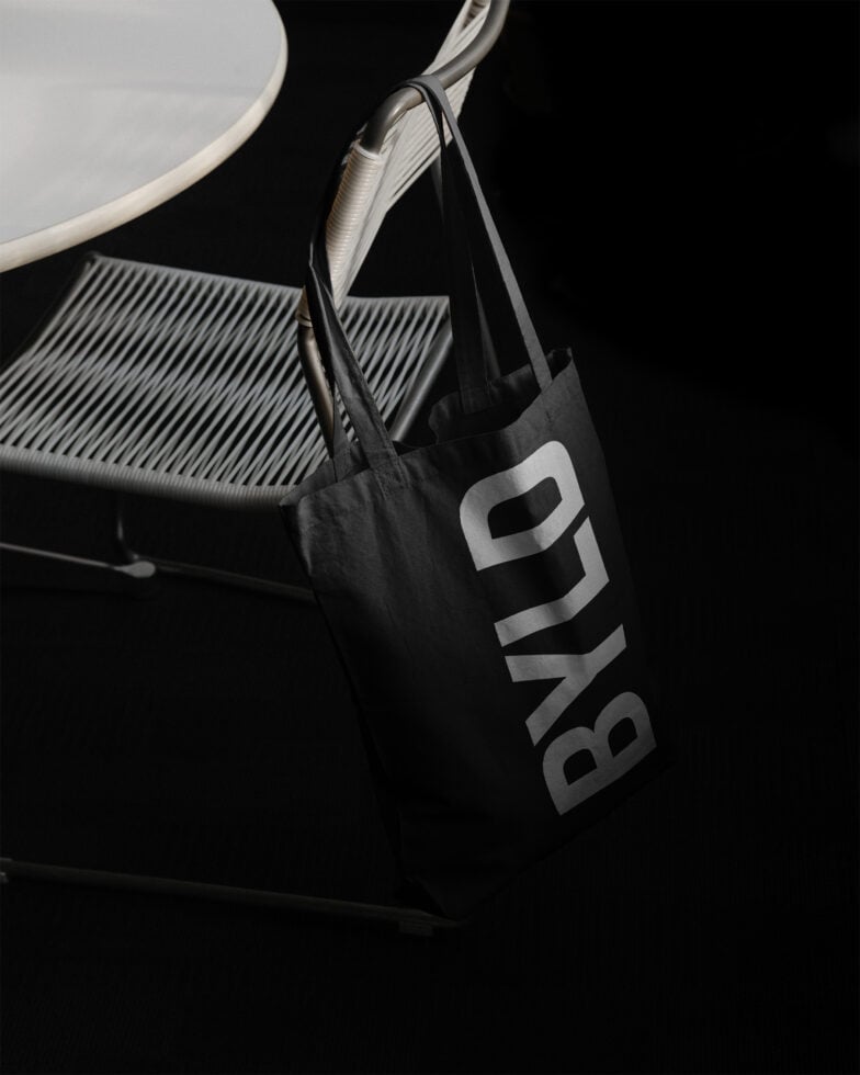 Black tote bag mockup hanging on a modern chair next to a round table, perfect for designers seeking realistic product presentation graphics.