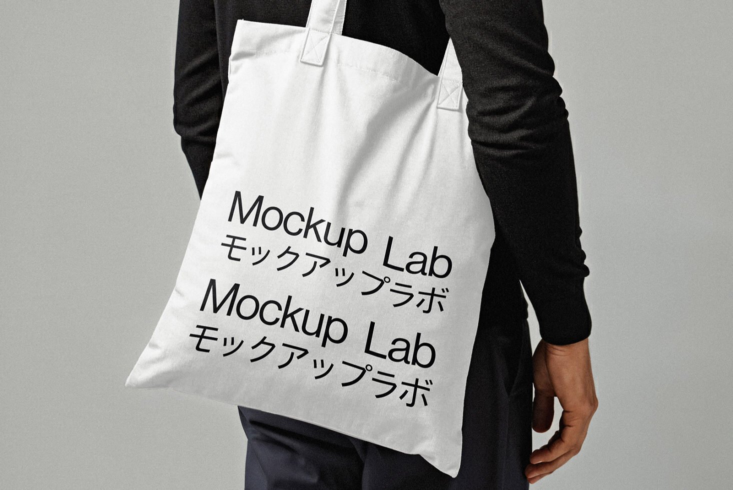 Tote bag mockup with black text Mockup Lab in English and Japanese on white fabric held by person in dark clothing design template for designers.
