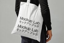 Tote bag mockup with black text Mockup Lab in English and Japanese on white fabric held by person in dark clothing design template for designers.