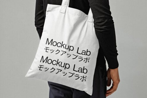 Tote bag mockup with black text Mockup Lab in English and Japanese on white fabric held by person in dark clothing design template for designers.