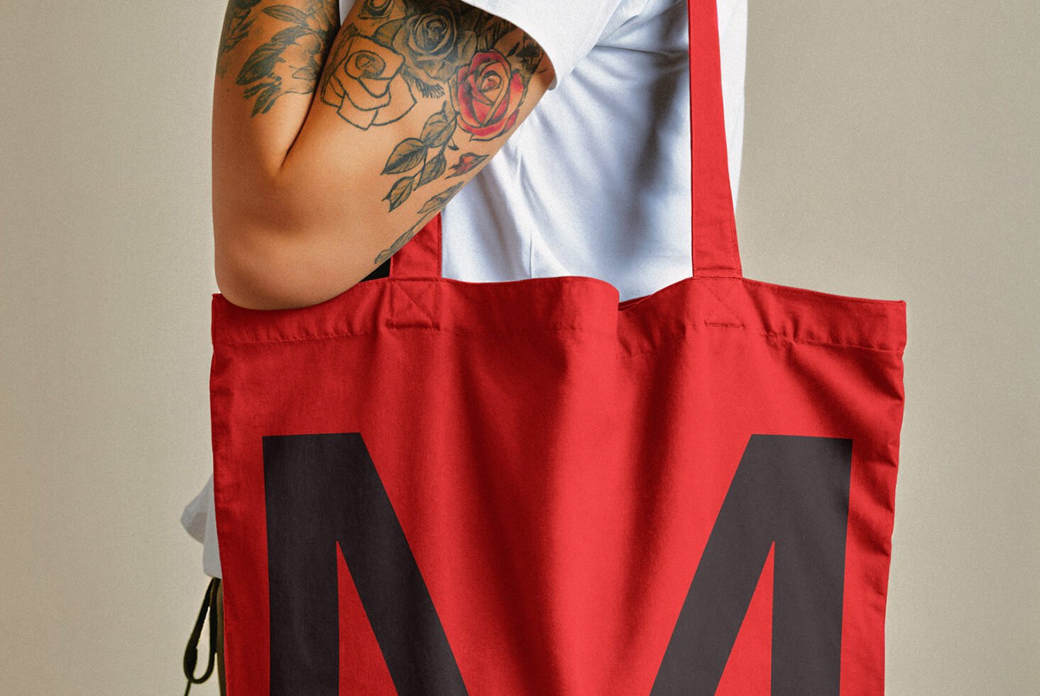 Red tote bag with bold black letter print held by a tattooed arm wearing a white shirt. Ideal for mockups, design assets, product templates.