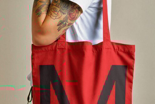 Red tote bag with bold black letter print held by a tattooed arm wearing a white shirt. Ideal for mockups, design assets, product templates.
