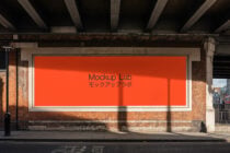 Street billboard mockup with solid orange background on brick wall under industrial bridge