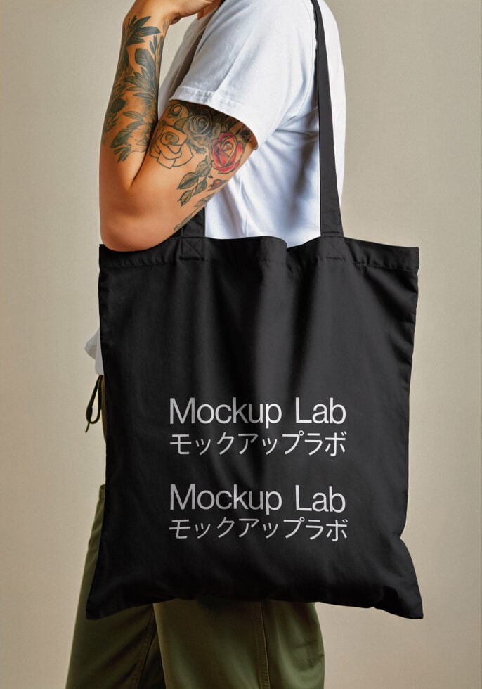 Tattooed person holding black tote bag with Mockup Lab logo. Ideal for tote bag mockup templates. Keywords: tote bag mockup, graphic design, template.