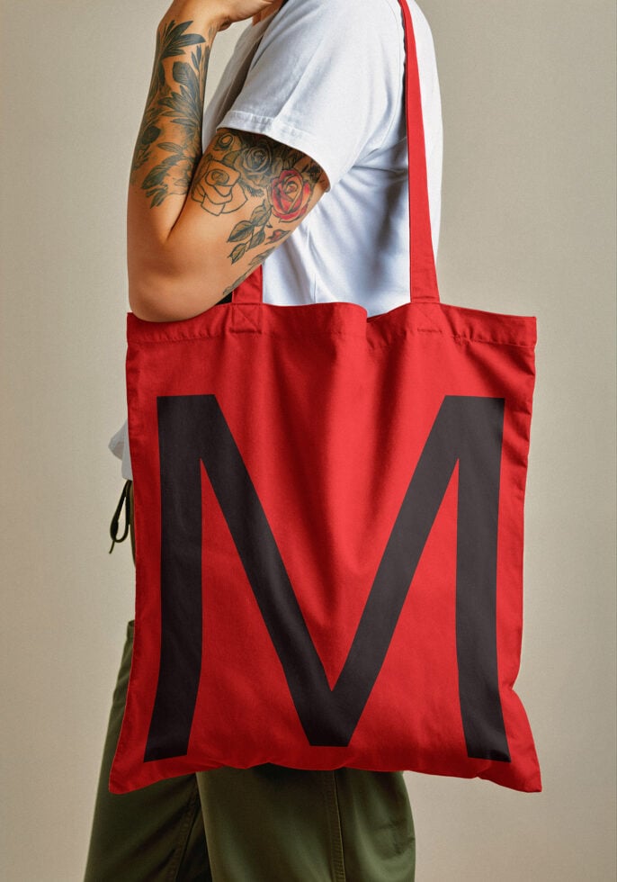 Red tote bag mockup with large black letter M, worn by tattooed person in white shirt; ideal for showcasing designs; eco-friendly fashion accessory