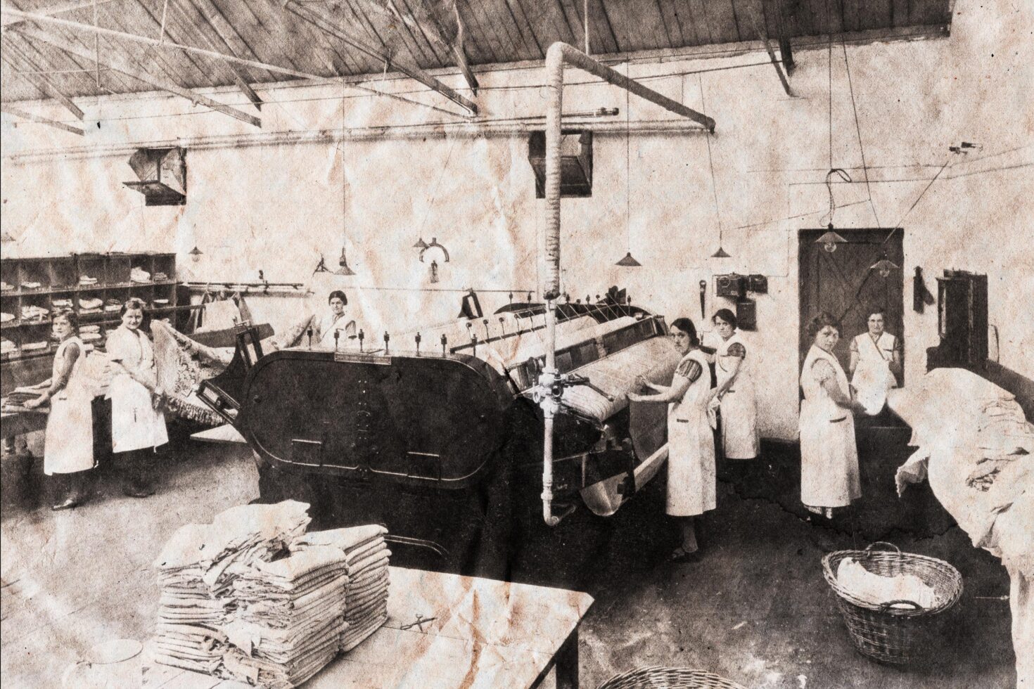 Vintage laundry workers in an industrial setting with large ironing machine and folded linens, perfect for designers CSS, retro graphics, nostalgic templates.