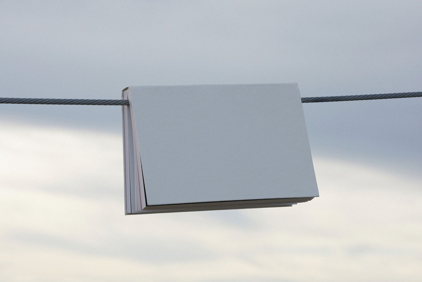 Minimalistic landscape mockup featuring a blank book hanging on a wire against a cloudy sky. Ideal for showcasing book covers and design projects.