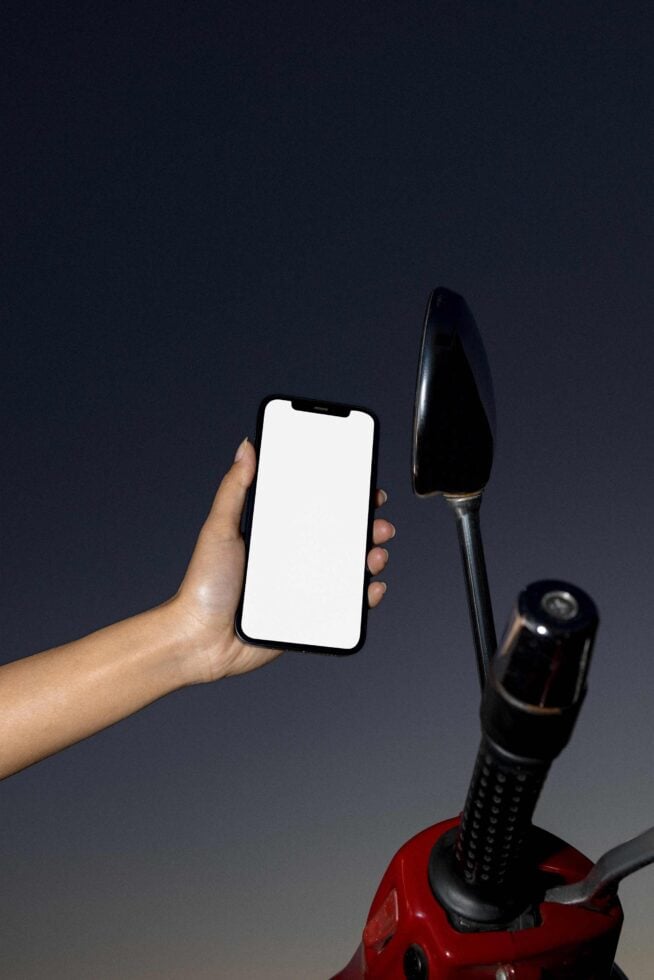 Mockup of a hand holding a smartphone with a blank screen beside a motorcycle handlebar against a dark background ideal for app design presentations