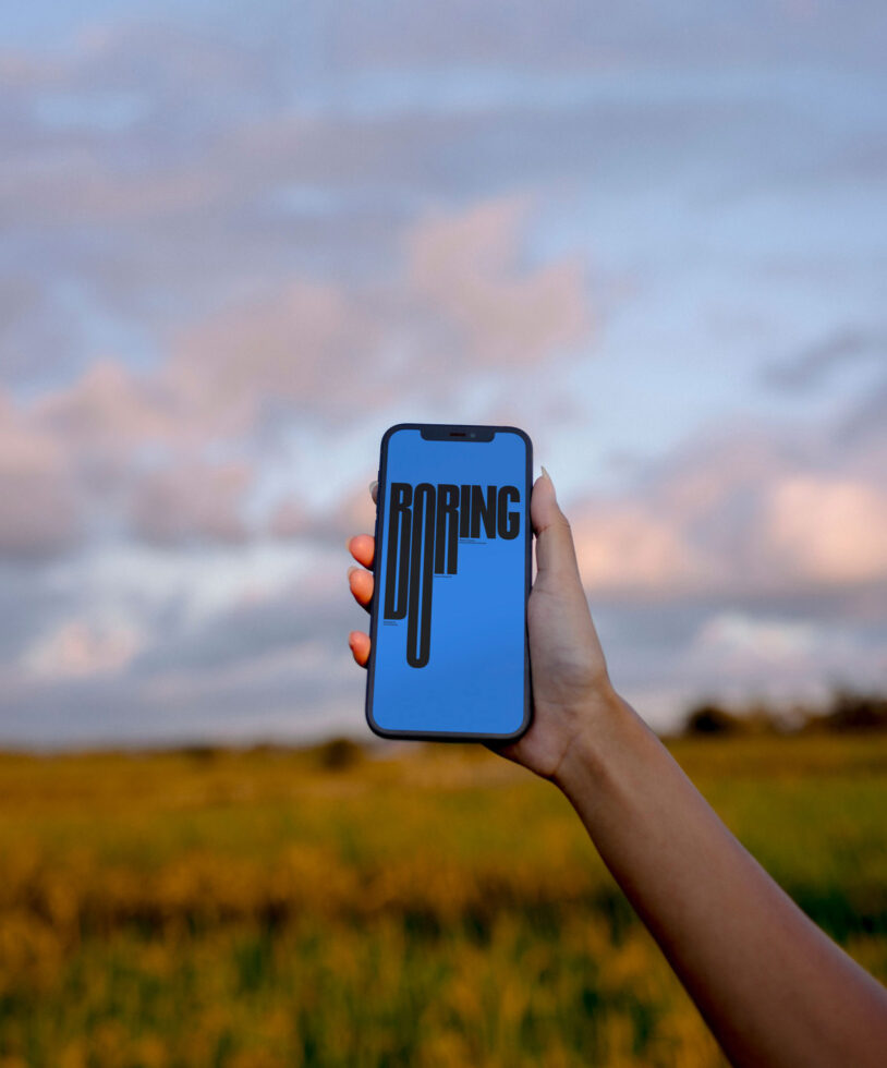Hand holding phone with bright blue screen displaying bold black BORING text in open field