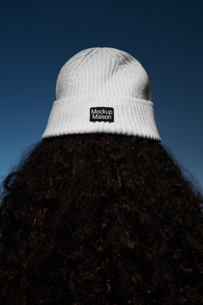 White beanie hat mockup with branding label on the back worn by person with curly hair against a blue sky. Ideal for fashion design templates.