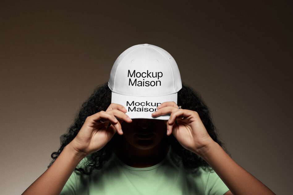 Person in green shirt holding white baseball cap in front of face on neutral brown backdrop