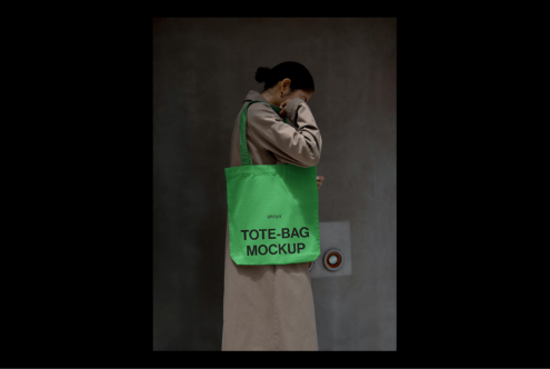Green tote bag mockup on shoulder of person in beige coat standing against gray wall. Perfect for design presentations and showcasing brand graphics.