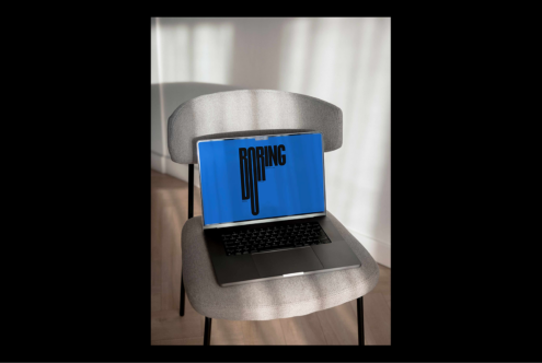 Laptop displaying bold BORING typography on blue background, set on a gray chair indoors. Perfect for designers seeking graphic inspiration.